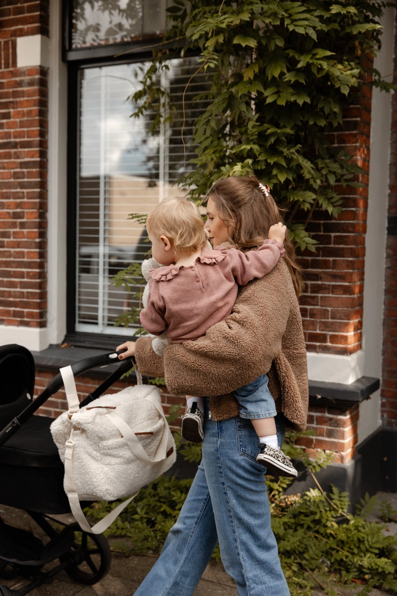 Moeder draagt kind terwijl teddy luiertas cream aan de kinderwagen hangt